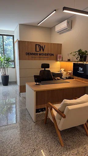 Recepção do consultório, com mesa de madeira, poltrona e painel com a marca Denner Weverton